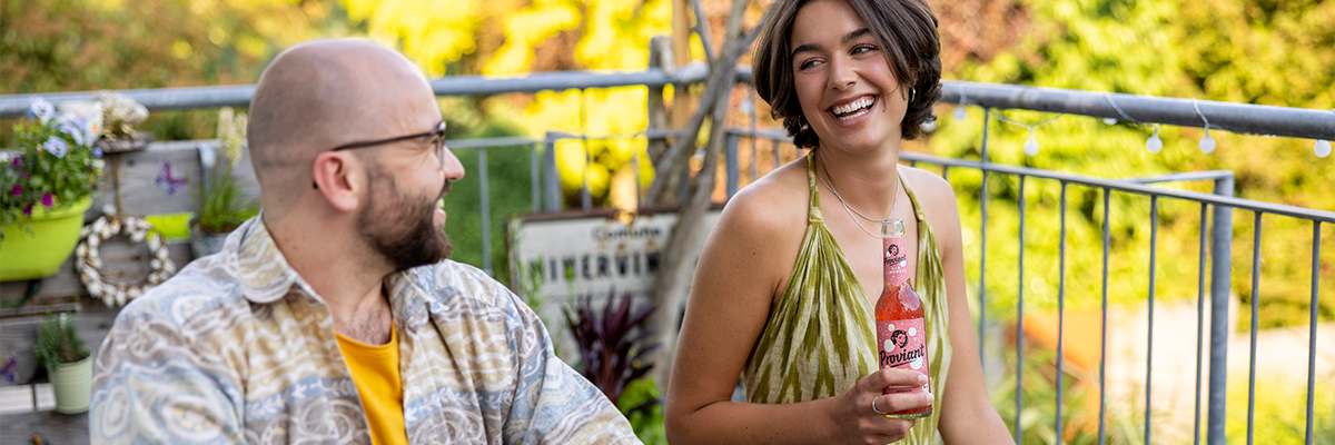 Zwei Personen sitzen auf einem Balkon, eine hält eine Flasche Proviant, im Hintergrund Pflanzen und Geländer.